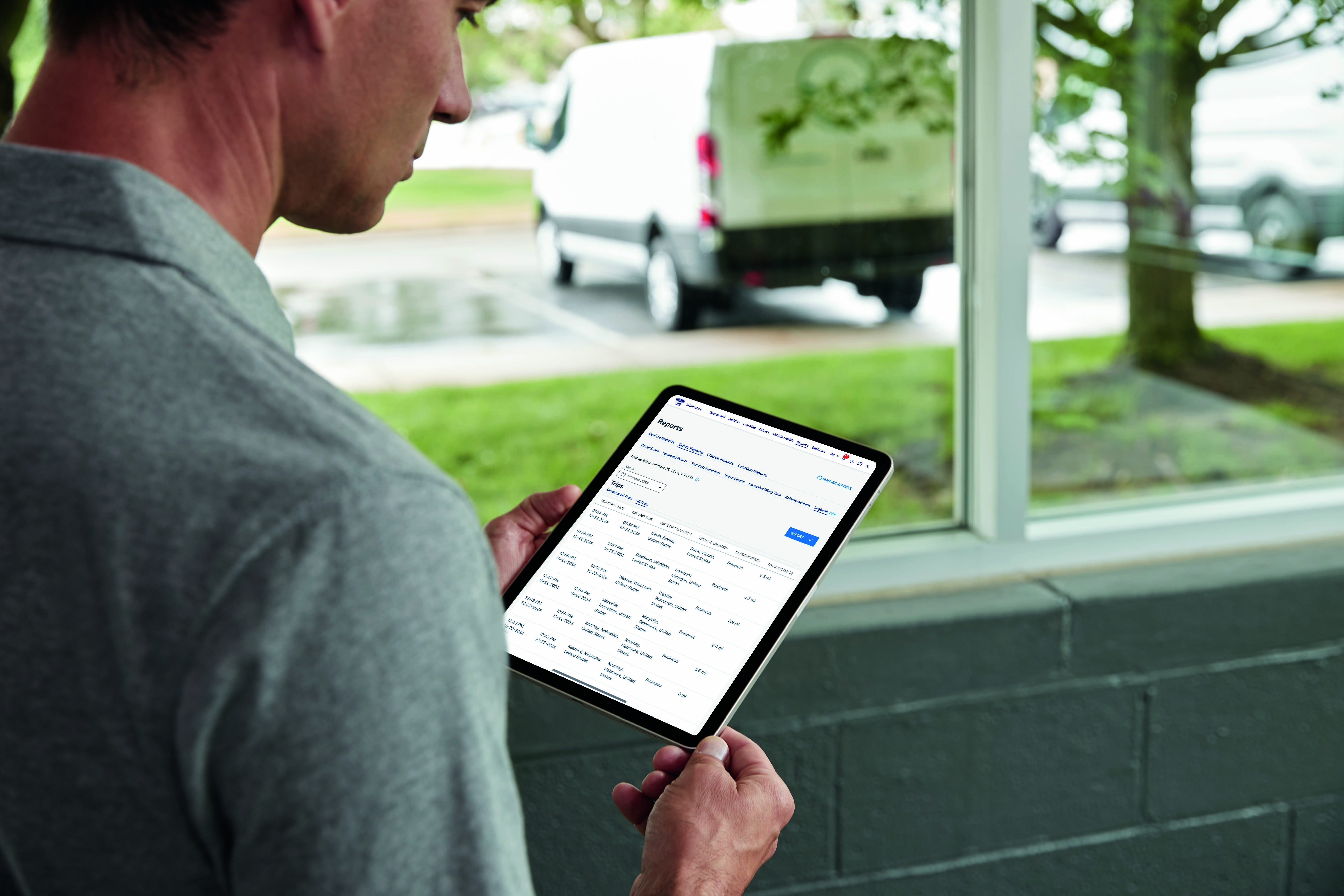 A man holds a tablet in front of a window. Outside the window, a van sits in the parking lot.